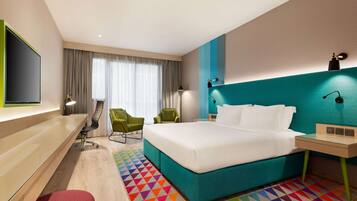 A modern hotel room with a large bed, two chairs, a desk, and a TV.