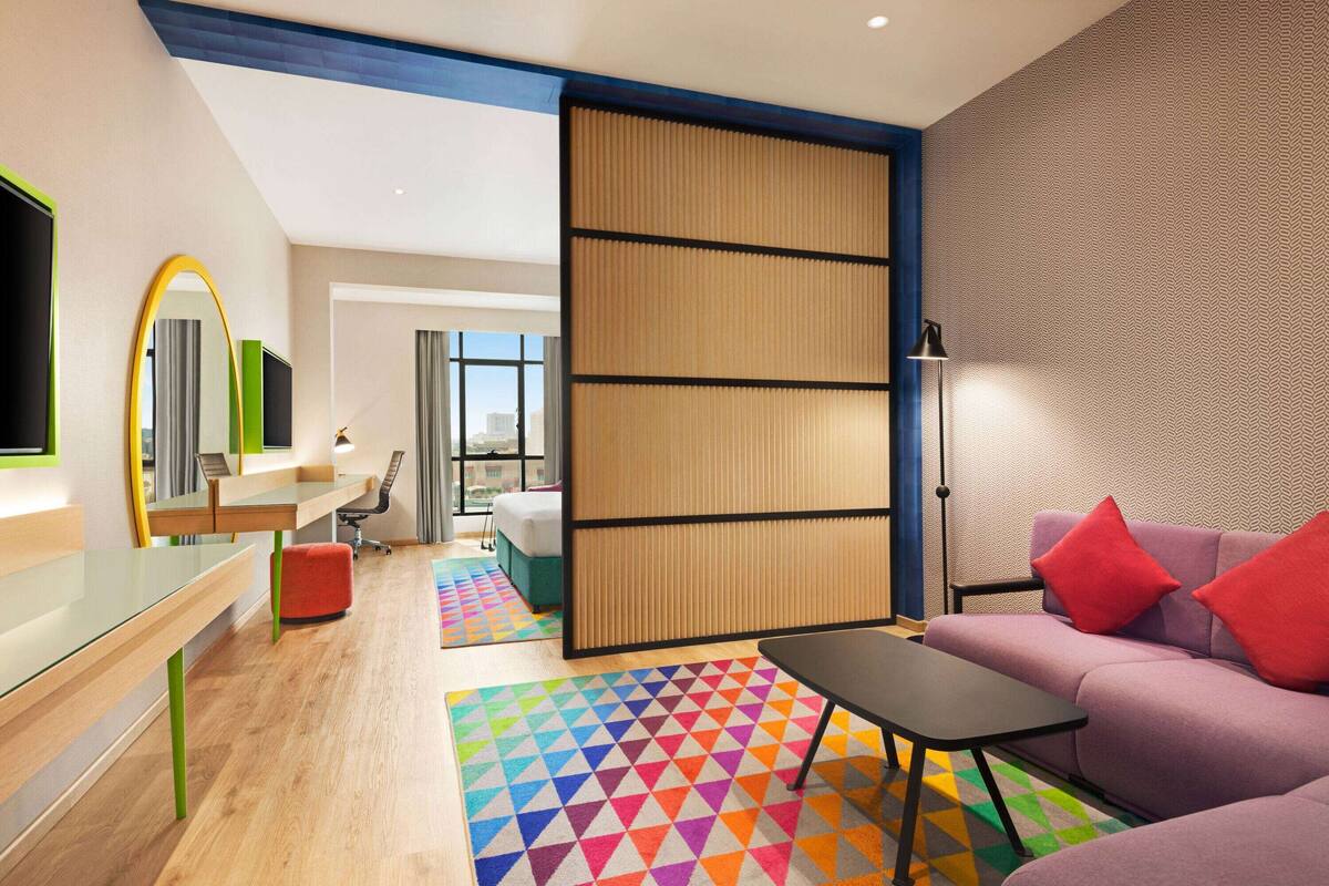 Una habitación de hotel moderna con un sofá grande, una mesita, un escritorio con una silla, una alfombra geométrica de colores y un ventanal con vistas a la ciudad.