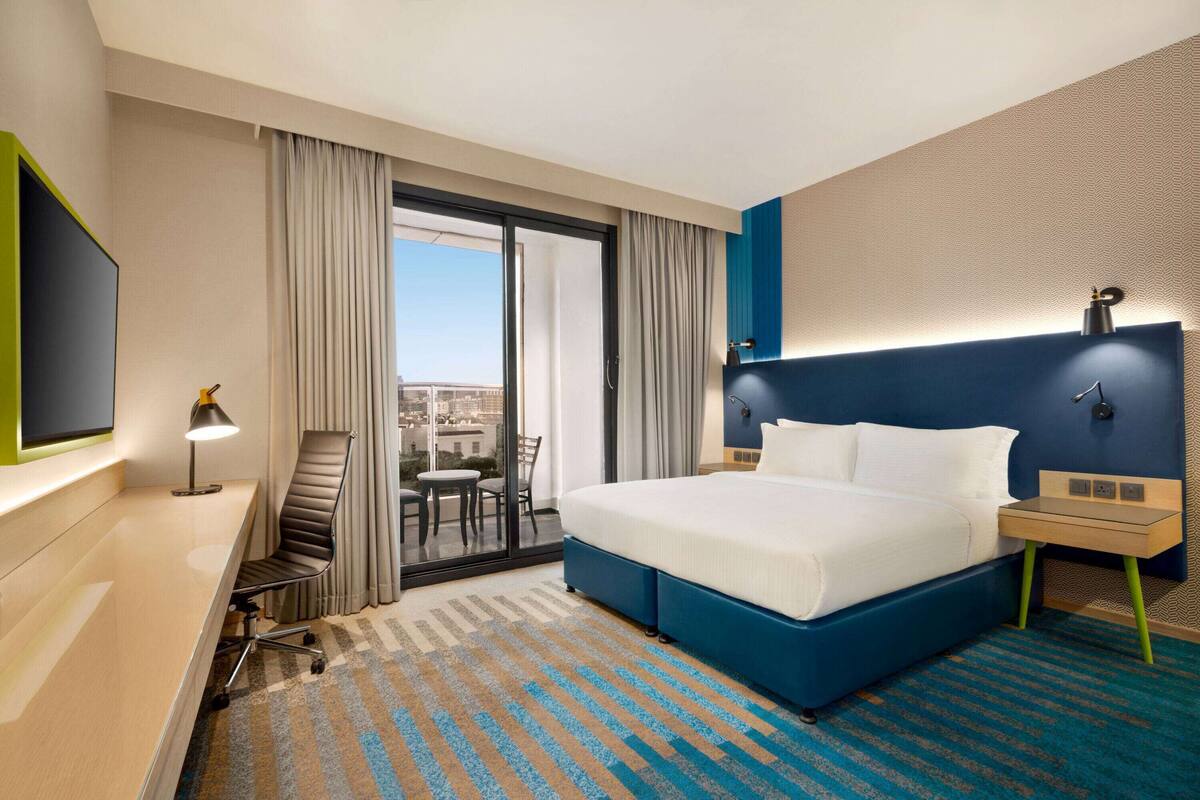 Habitación de hotel con una cama grande, un escritorio, una silla, un televisor y vistas a la ciudad a través del ventanal.