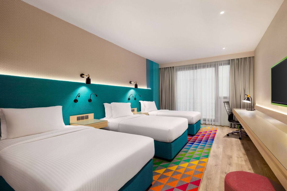 Habitación de hotel con tres camas, un escritorio, un televisor y una alfombra colorida.
