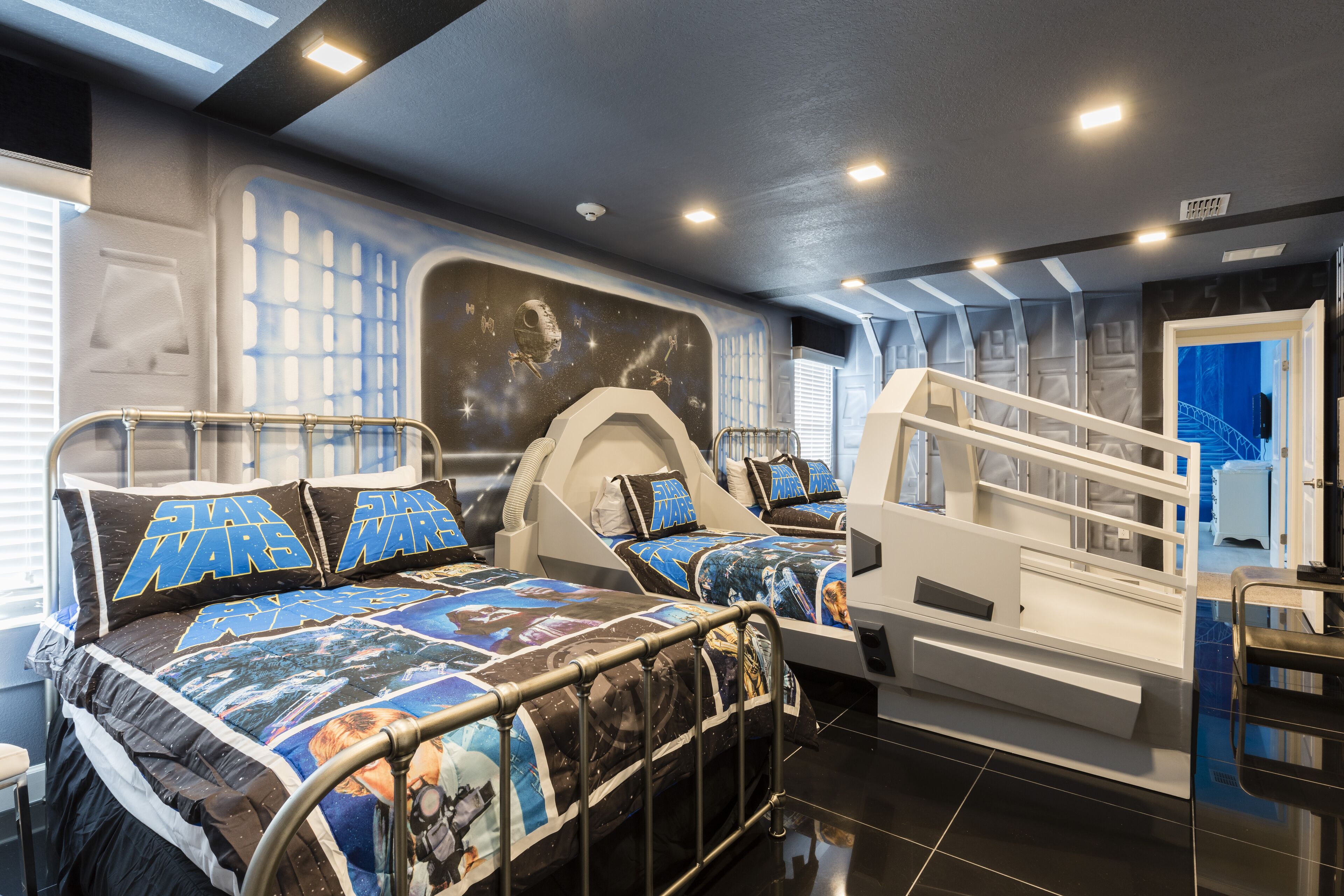 A Star Wars-themed bedroom with two beds, each with matching Star Wars bedding and pillows. The room features a unique design with a space-themed wall and a staircase leading to an upper level.