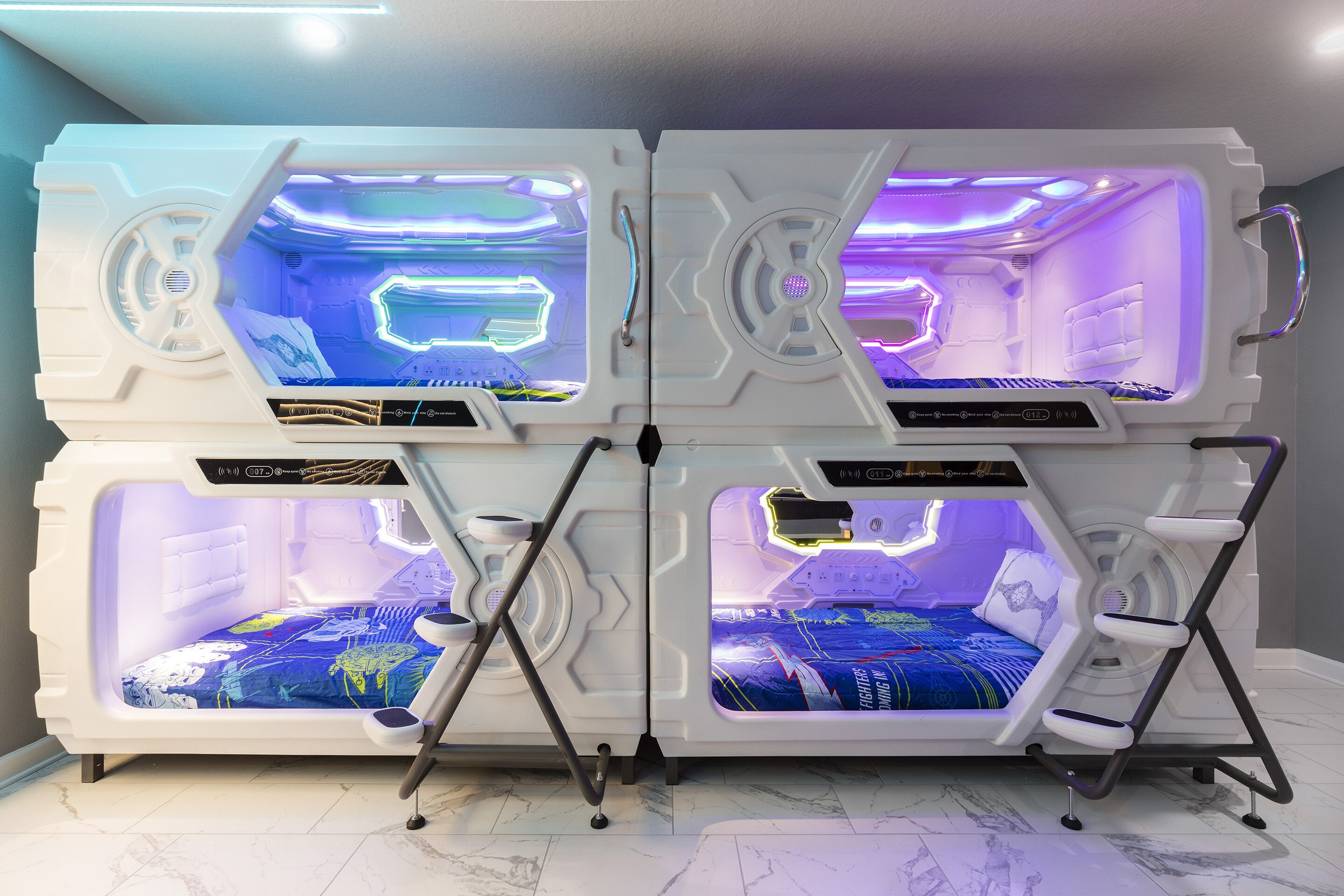 Vier futuristische, verlichte pods met bedieningspanelen, elk met een bed en een voetenbankje.