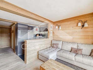 A modern living room with a grey sofa, wooden walls, and a kitchen area visible through a glass door. - Sunny apartment for 6, ski-in/ski-out in Méribel (Méribel)