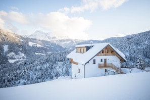 Una casa di montagna ricoperta di neve con facciata in legno, un balcone e vista su alberi e montagne innevate.