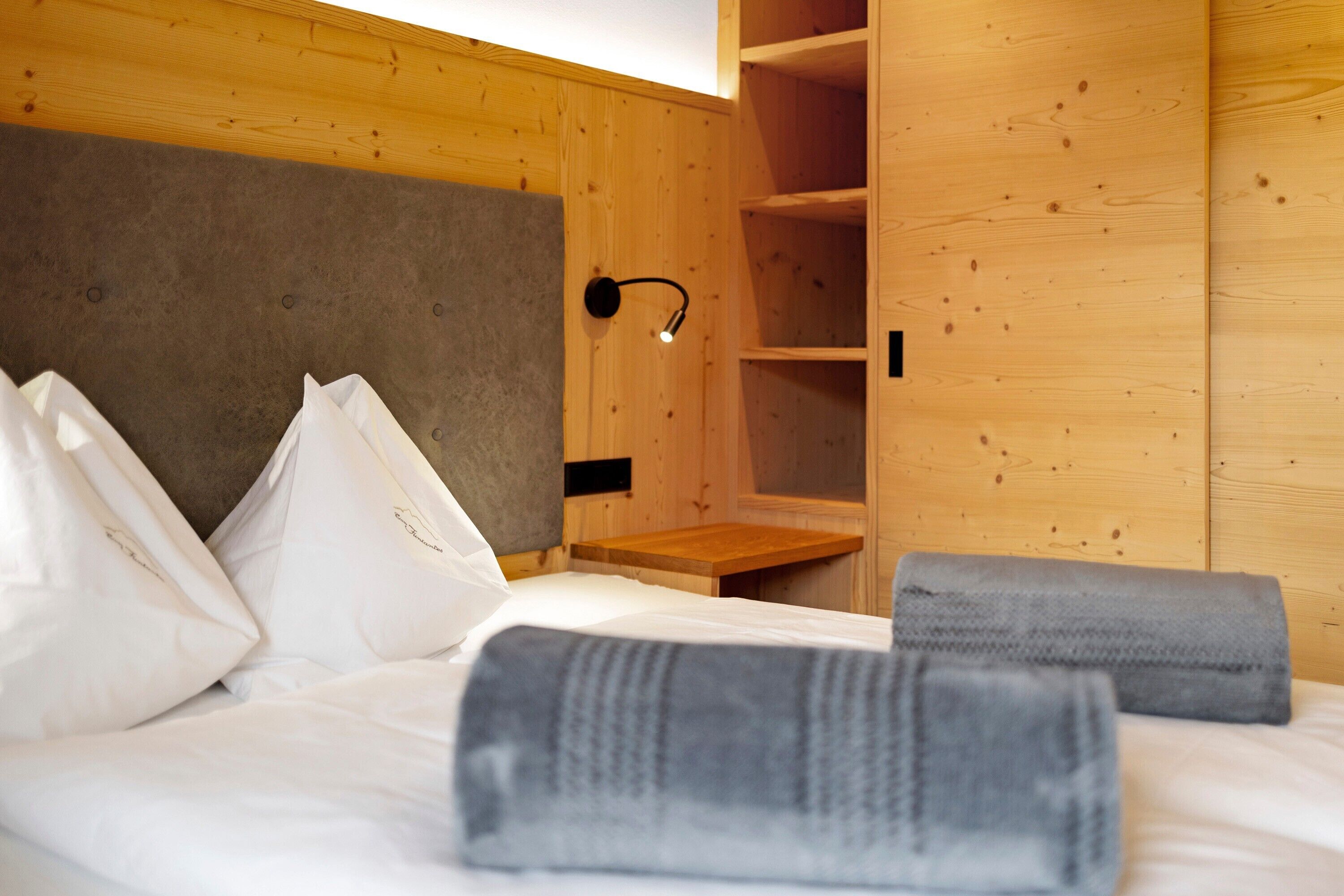 Una habitación de madera con una cama, almohadas, una mesita de noche y una lámpara fijada en la pared.