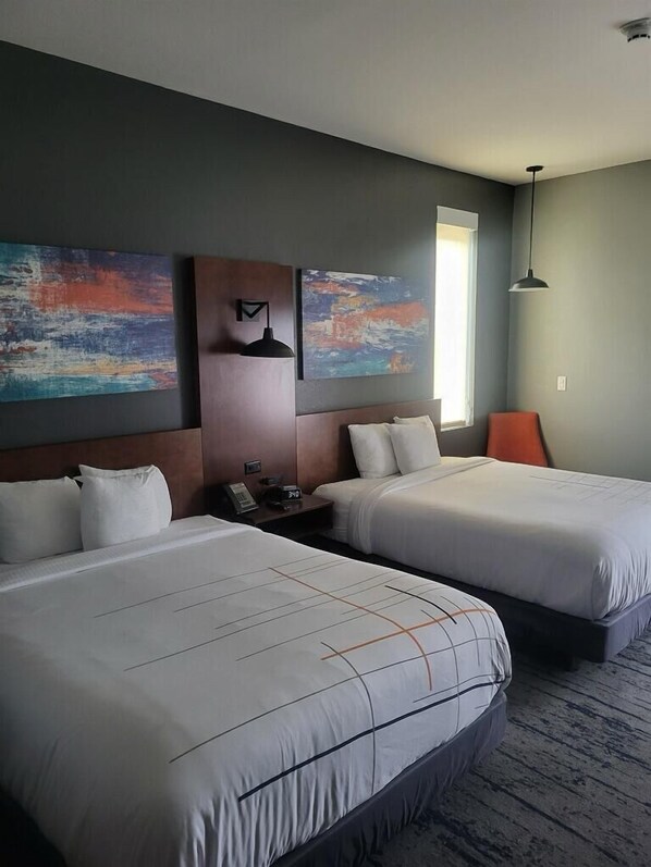 A hotel room with two beds, a nightstand, a wall-mounted lamp, and abstract paintings.