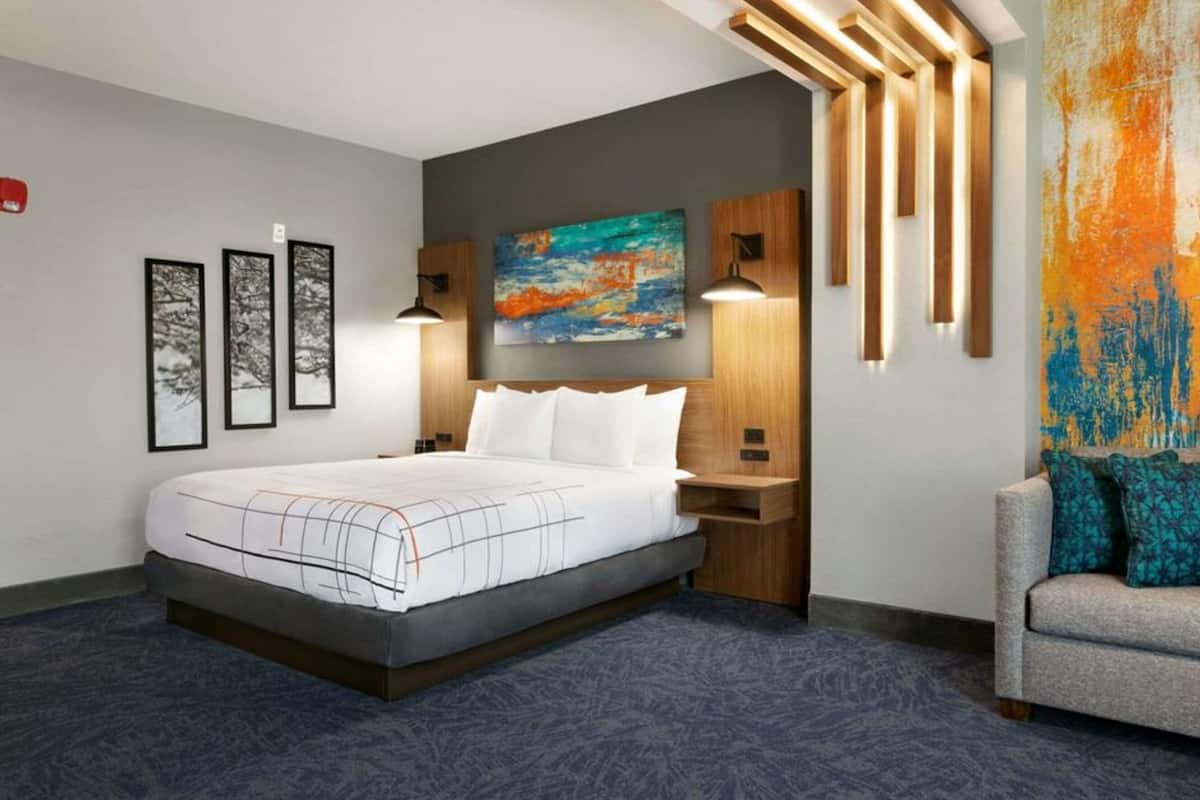 Une chambre d’hôtel moderne avec un grand lit, un bureau et un canapé. Les murs sont ornés de tableaux encadrés et, au-dessus du lit, on peut admirer une peinture abstraite aux couleurs vives.