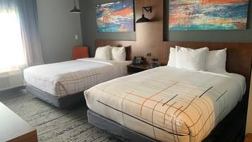 Habitación de hotel con dos camas, un escritorio, una silla, una lámpara y obras de arte en las paredes.