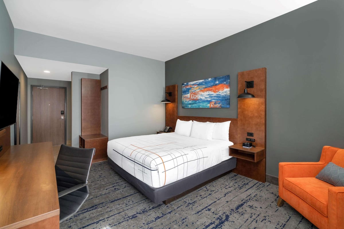 Une chambre d’hôtel moderne avec un grand lit, un bureau en bois, un fauteuil orange et un tableau au mur.