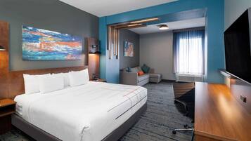 A hotel room with a large bed, a desk, a sofa, and a TV. There are paintings on the walls and a window with curtains.