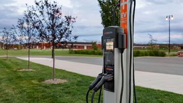 Uma estação de carregamento de veículos elétricos pública com visor digital, situada numa área ajardinada com uma estrada e edifícios ao fundo.
