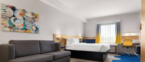 Una habitación de hotel moderna con un sofá gris, una cama con cabecera blanca, un escritorio con computadora y un cuadro abstracto y colorido en la pared.