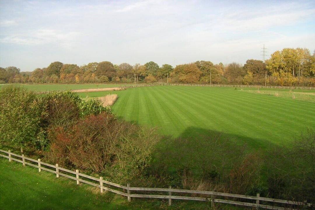 A large, open grassy field with a wooden fence in the foreground and trees in the background.