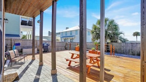 A covered deck with picnic tables, a barbecue grill, and a poolside area with a wooden fence and palm trees.
