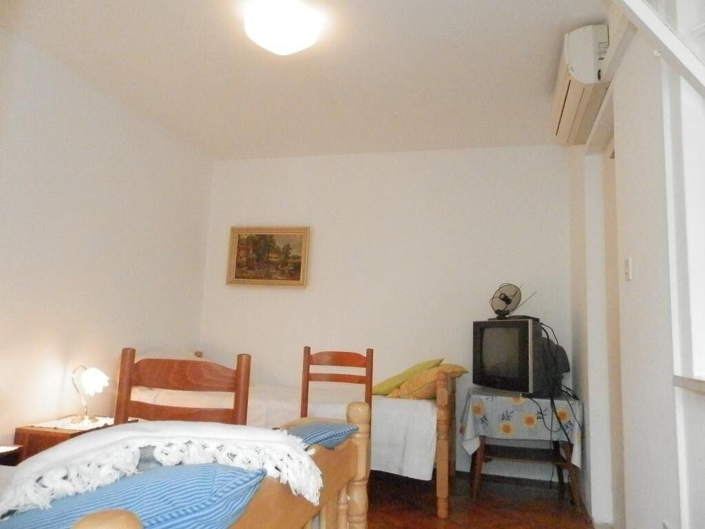 A small, simple room with two wooden beds, a single chair, a small table with a television, and a painting on the wall.