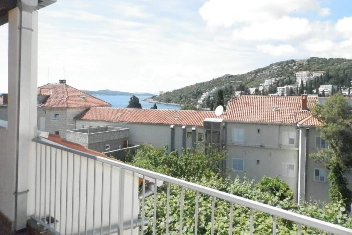 A balcony with a view of a coastal town, featuring terraced houses with red roofs and a body of water in the distance.