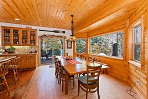A wooden cabin interior with a dining area, kitchen, and large windows. - Bearadise Found by Destination Big Bear : Luxury Log Home in Upper Moonridge! Game Room with Foosball & Shuffleboard! (Big Bear City)