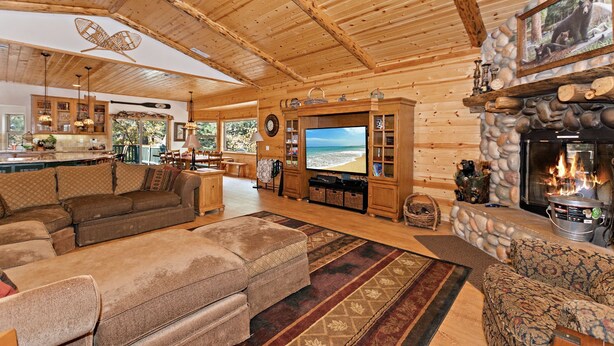 Acogedor interior de cabaña de madera con televisor grande, chimenea y comedor con vistas al exterior.