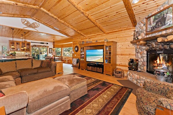 A log cabin interior with a large living area, fireplace, stone wall, and a television displaying a beach scene.