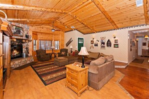 A cozy cabin interior with a stone fireplace, wooden ceiling, and a living area furnished with a sofa, armchair, and a wooden side table. - Bearadise Found by Destination Big Bear : Luxury Log Home in Upper Moonridge! Game Room with Foosball & Shuffleboard! (Big Bear City)