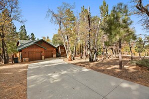 A wooden cabin with a garage, surrounded by trees and a clear sky. - Bearadise Found by Destination Big Bear : Luxury Log Home in Upper Moonridge! Game Room with Foosball & Shuffleboard! (Big Bear City)