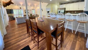 A spacious dining area with a wooden table and chairs, a kitchen in the background, and a mounted deer head on the wall.