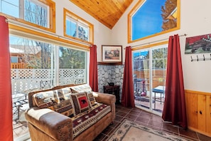 Um quarto acolhedor com lareira de pedra, sofá de couro com almofadas estampadas e grandes janelas com vista para o exterior.