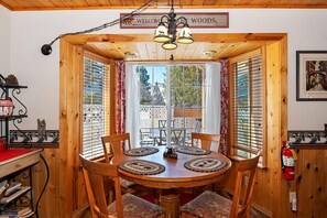 A wooden dining area with a round table and chairs, a chandelier, and a view of the outdoors through a window with blinds.