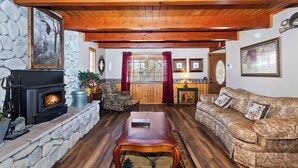 A cozy living room with a stone fireplace, wooden ceiling beams, a sofa, a coffee table, and a stone wall. - Nestled in the Upper Moonridge with Beautiful Views! Outdoor Hot Tub in Fenced Yard! Game Room! Two Master Suites! (Big Bear City)