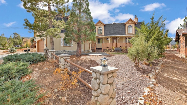 A two-story house with a stone fence, a front porch, and a driveway. - Nestled in the Upper Moonridge with Beautiful Views! Outdoor Hot Tub in Fenced Yard! Game Room! Two Master Suites! (Big Bear City)