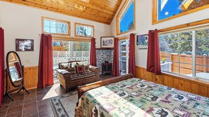 A spacious room with a large bed, wooden ceiling, multiple windows, and a stone fireplace. - Nestled in the Upper Moonridge with Beautiful Views! Outdoor Hot Tub in Fenced Yard! Game Room! Two Master Suites! (Big Bear City)