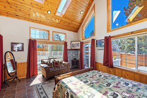 A spacious room with a large bed, wooden ceiling, multiple windows, and a stone fireplace.