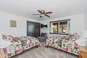A room with two beds, a ceiling fan, a window with curtains, a door, and a small gray chair. - Village Retreat by Destination Big Bear : Custom Build, New Construction! Pool Table! Shuffleboard! Near Village! (Big Bear Lake)