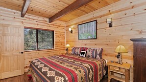 Een slaapkamer in een houten blokhut met een bed, twee bedlampen, een raam met zonwering, een lijst met een afbeelding aan de muur en een houten nachtkastje.