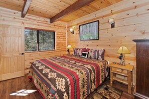 A log cabin bedroom with a bed, two bedside lamps, a window with blinds, and a framed picture on the wall.