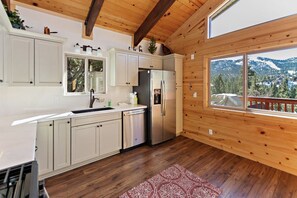 A modern kitchen with wooden beams, white cabinets, stainless steel appliances, and a large window offering a mountain view.