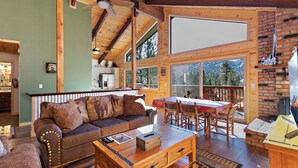 A cozy cabin interior with a wooden dining table, a sofa with animal-themed cushions, and a large window offering a view of the outdoors.