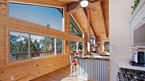 Interieur van een blokhut met houten wanden, grote ramen, een plafondventilator, een keukenhoek en uitzicht op een besneeuwde berg.