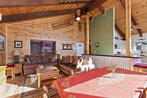 Interior acogedor de una cabaña con techo de madera, un comedor con mantel rojo y una cocina visible a través de una puerta abierta.