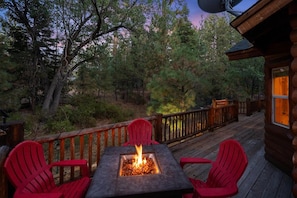 A deck with a fire pit table and red chairs, surrounded by a wooden deck and trees.