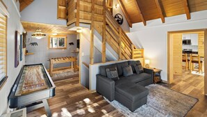 A spacious living area with a wooden staircase, a foosball table, a sofa, and a kitchen area. - Mooncrest Manor by Destination Big Bear : Expansive Mountain Views! Large Family Spaces! Pet Friendly! Game Room! (Big Bear Lake)