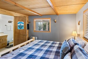 A bedroom with a checkered bedspread, a wooden headboard, a window with blinds, a door with a sign, and a small bedside table with a lamp. - Mooncrest Manor by Destination Big Bear : Expansive Mountain Views! Large Family Spaces! Pet Friendly! Game Room! (Big Bear Lake)