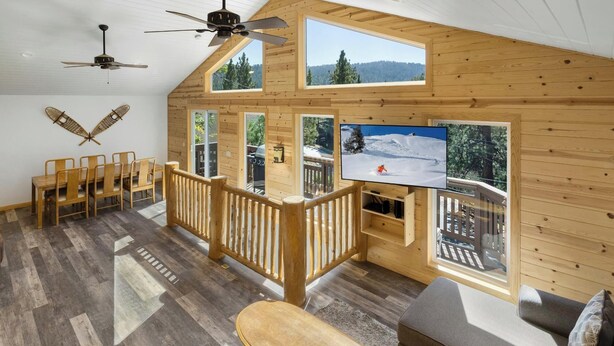A wooden cabin interior with a dining area, a sofa, a TV, and a balcony with a ski view. - Mooncrest Manor by Destination Big Bear : Expansive Mountain Views! Large Family Spaces! Pet Friendly! Game Room! (Big Bear Lake)