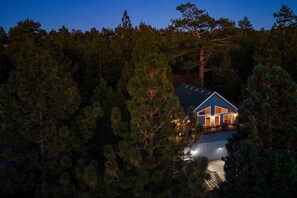 A house surrounded by dense forest at dusk. - Mooncrest Manor by Destination Big Bear : Expansive Mountain Views! Large Family Spaces! Pet Friendly! Game Room! (Big Bear Lake)