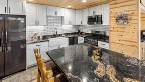 A modern kitchen with white cabinets, a black marble countertop, stainless steel appliances, and a wooden ceiling. - Mooncrest Manor by Destination Big Bear : Expansive Mountain Views! Large Family Spaces! Pet Friendly! Game Room! (Big Bear Lake)