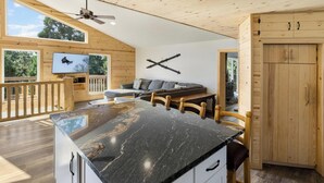 A modern kitchen with a marble countertop, wooden cabinets, and a dining area with a wooden table and chairs. - Mooncrest Manor by Destination Big Bear : Expansive Mountain Views! Large Family Spaces! Pet Friendly! Game Room! (Big Bear Lake)
