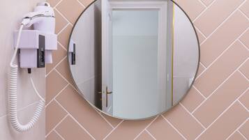 Un baño con un espejo redondo, un lavabo blanco, un grifo dorado, una planta en maceta y un secador de pelo en la pared.