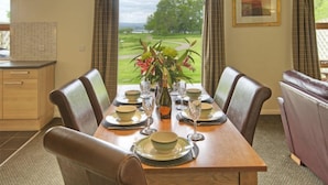 A dining area with a table set for four, a vase with flowers, and a view of a green landscape through large windows. - Drumlanrig Lodge with hot tub and views of Loch Lomond (Loch Lomond)