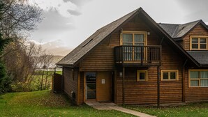 A two-story wooden house with a balcony on the upper floor, a front door, and a small porch area. - Cawdor  Diamond  Lodge with hot tub and Loch views (Balmaha)