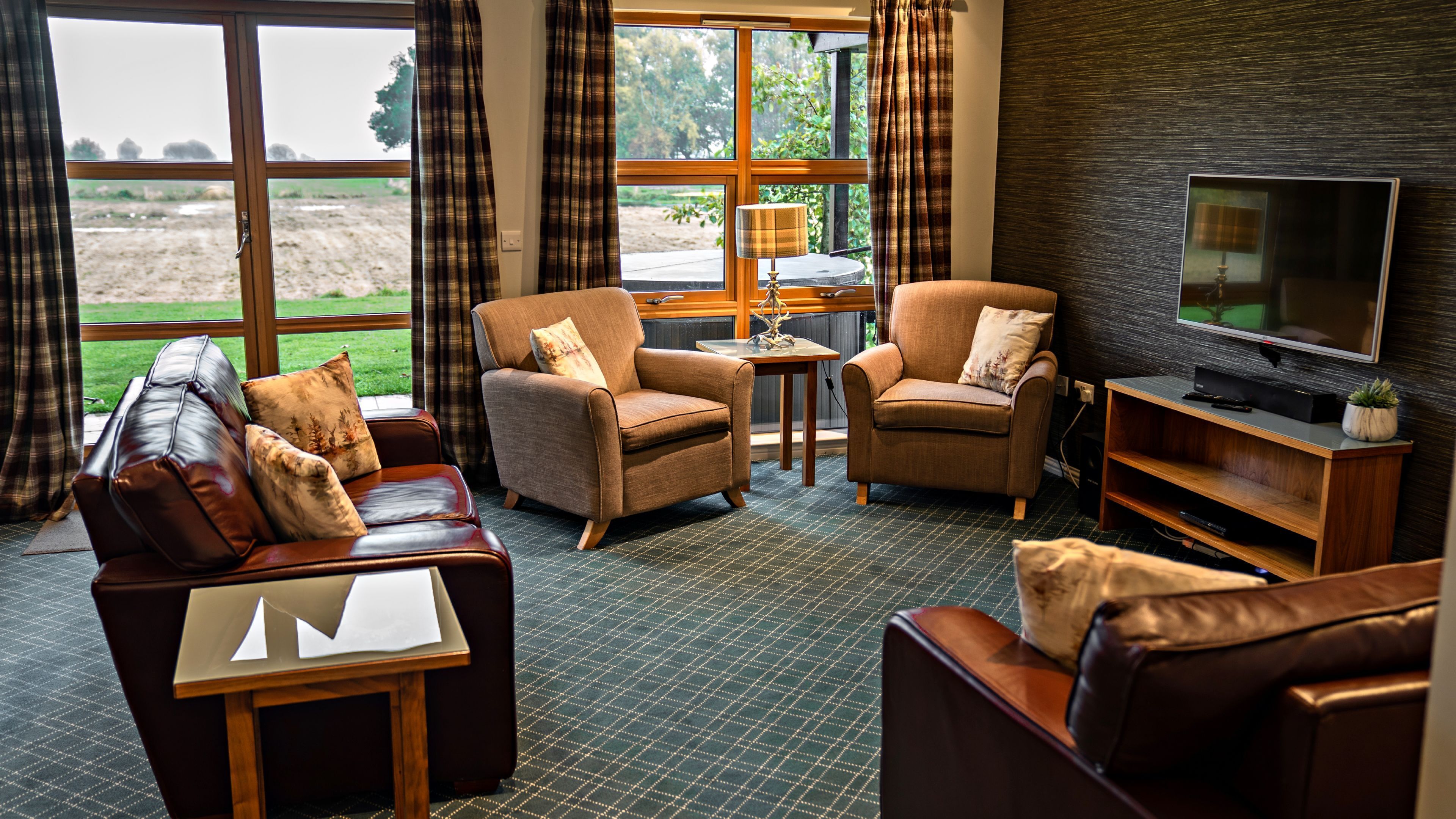A living room with leather sofas, armchairs, a coffee table, and a TV. Large windows offer a view of the outdoors.